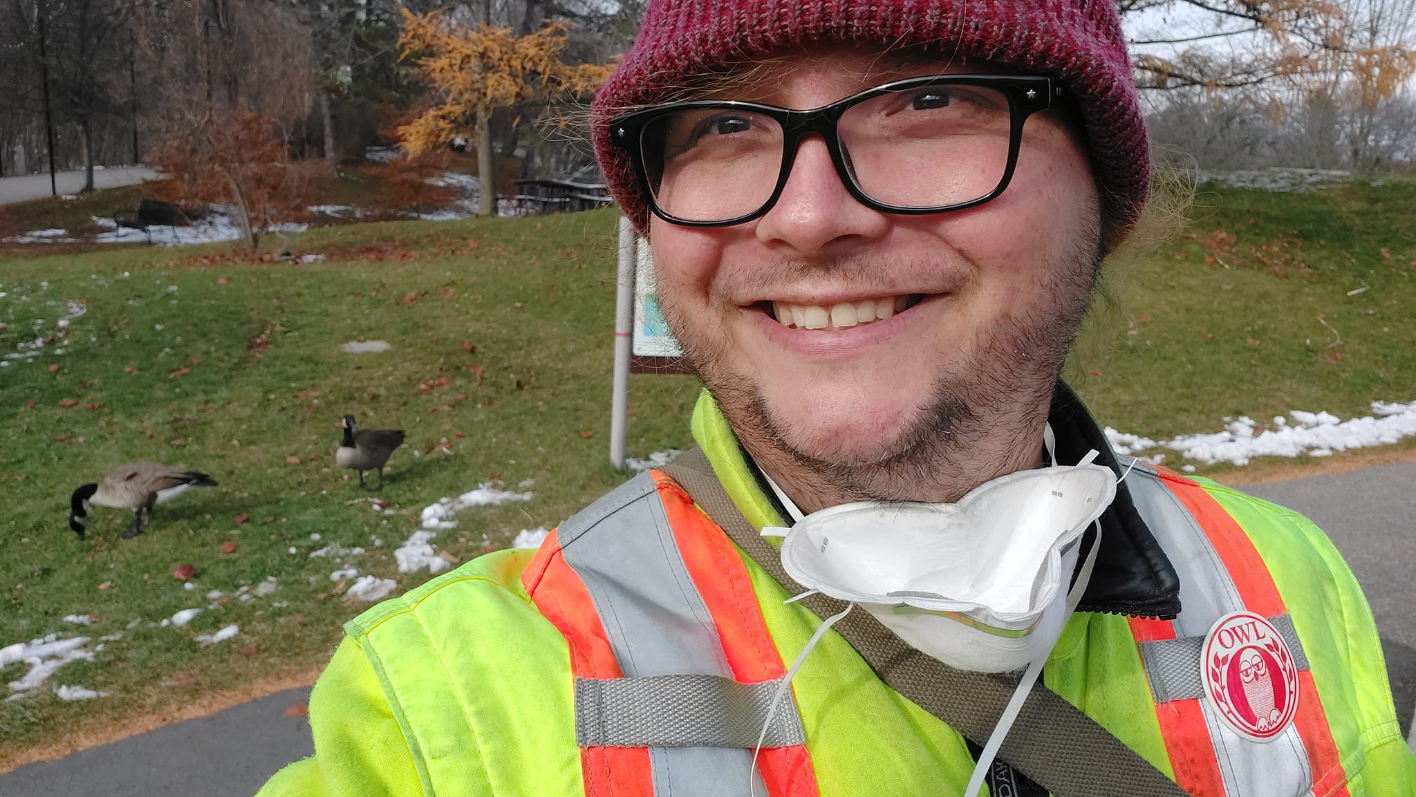 A man wearing a red toque, a high visibility winter coat and an N95 mask around his neck stands in front of some Canada geese on some grass